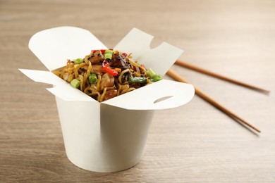Box of wok noodles with vegetables, meat and chopsticks on wooden table, closeup