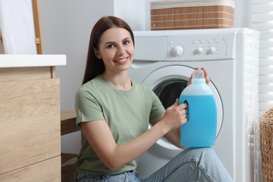 Photo of Woman sitting on floor near washing machine and holding fabric softener in bathroom, space for text