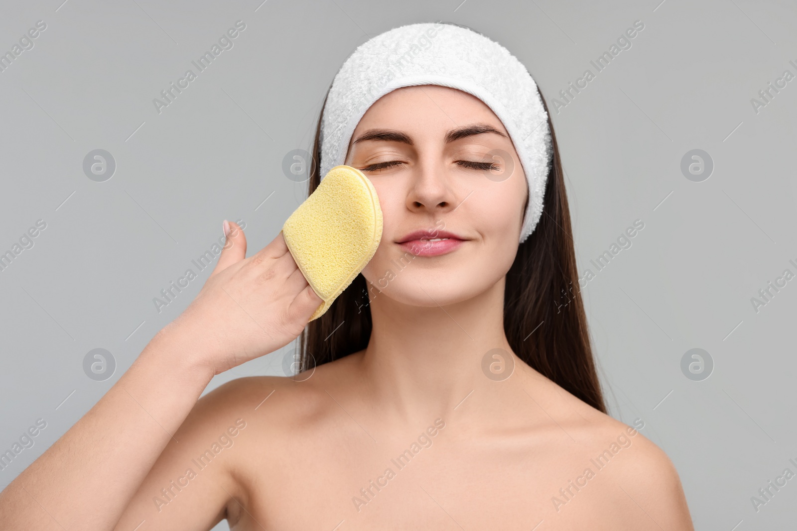 Photo of Young woman with headband washing her face using sponge on light grey background