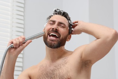Photo of Happy man with showerhead singing and washing his hair with shampoo indoors
