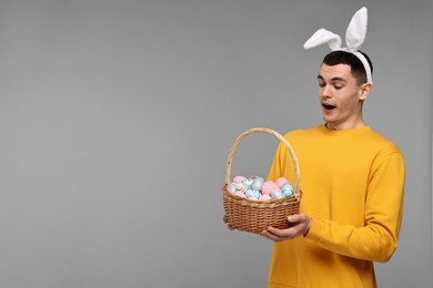 Photo of Easter celebration. Handsome young man with bunny ears holding basket of painted eggs on grey background. Space for text