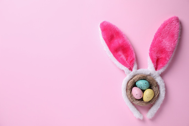 Headband with Easter bunny ears and dyed eggs in nest on pink background, flat lay. Space for text