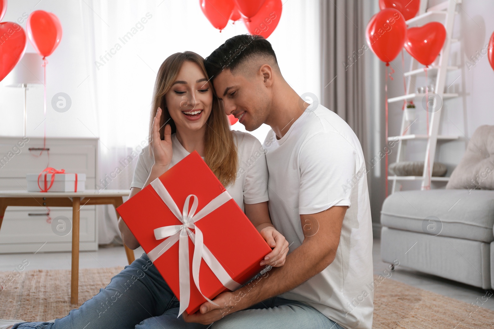 Photo of Man presenting gift to his girlfriend in room decorated with heart shaped balloons. Valentine's day celebration