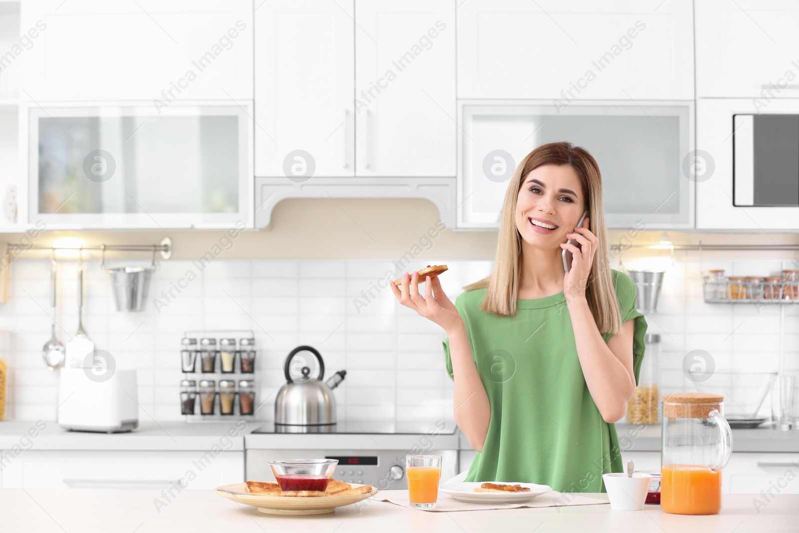 Photo of Beautiful woman eating tasty toasted bread with jam while talking on mobile phone at table in kitchen