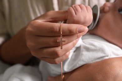 Mother holding Christian cross near newborn baby indoors, focus on hand