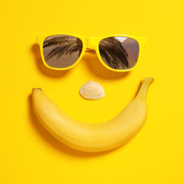 Funny face of sunglasses with reflection of palm trees, banana and seashell on yellow background, top view.
