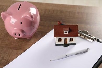 Photo of House model, piggy bank, clipboard and pen on wooden table