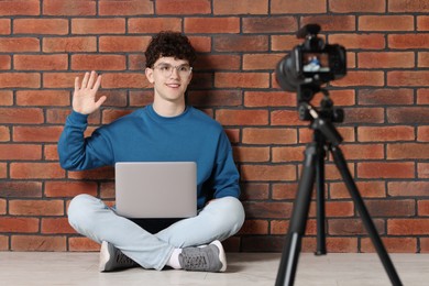 Smiling teenage blogger with laptop waving hello to his subscribers while streaming indoors