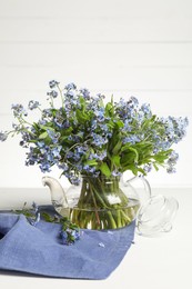 Bouquet of beautiful forget-me-not flowers in glass teapot and blue cloth on white table