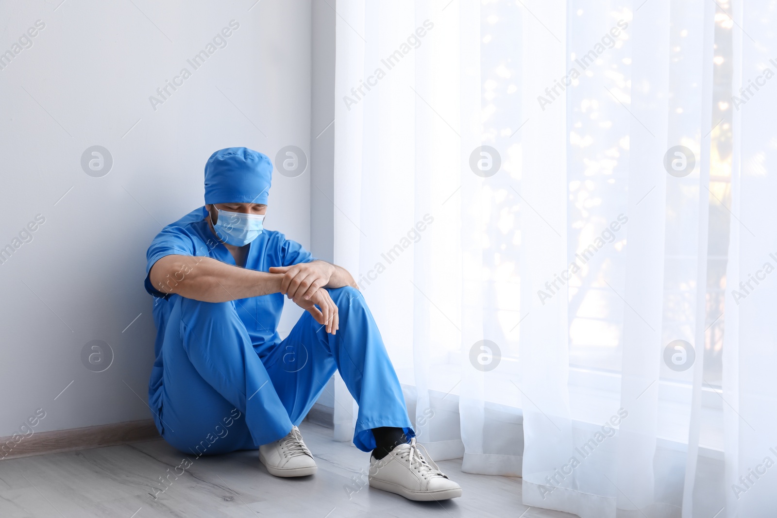 Photo of Exhausted doctor sitting on floor indoors, space for text. Stress of health care workers during COVID-19 pandemic