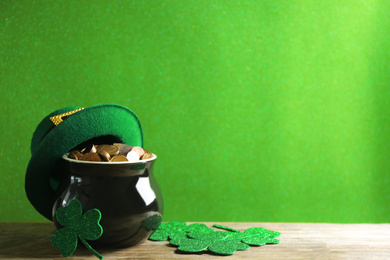 Photo of Pot of gold coins, hat and clover leaves on wooden table against green background, space for text. St. Patrick's Day celebration