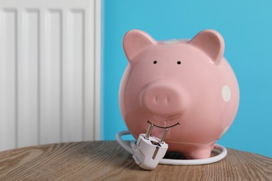 Photo of Piggy bank and plug on wooden table near heating radiator, space for text