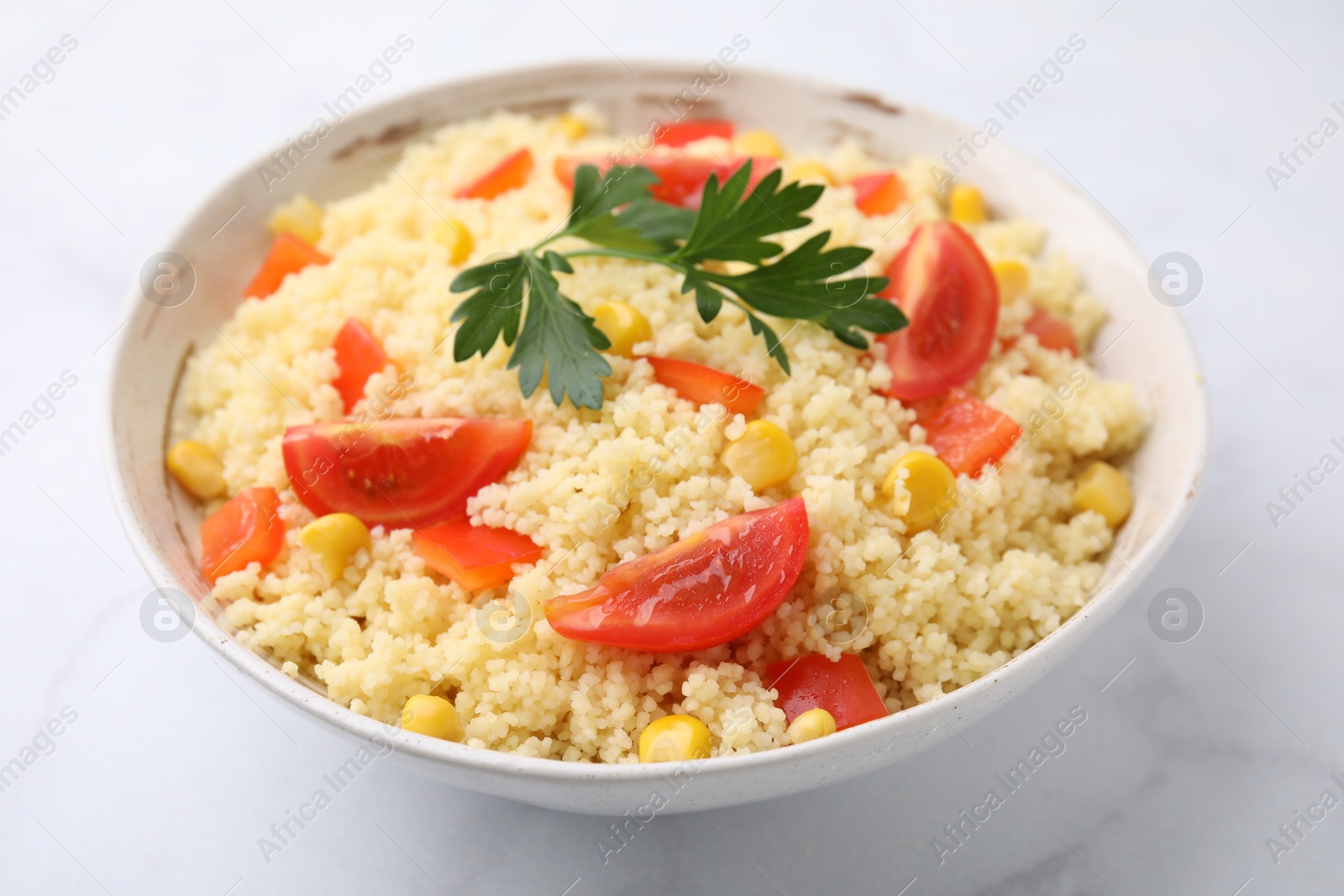 Photo of Tasty couscous with parsley, corn and tomatoes in bowl on white marble table, closeup