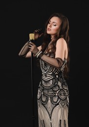 Beautiful young woman in stylish dress with microphone singing on black background, space for text