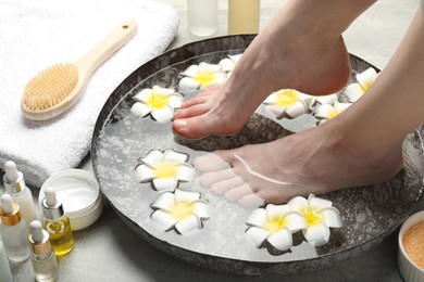 Photo of Woman soaking her feet in bowl with water and flowers on light grey floor, closeup. Spa treatment