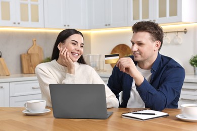 Happy couple with laptop and credit card shopping online at wooden table in kitchen