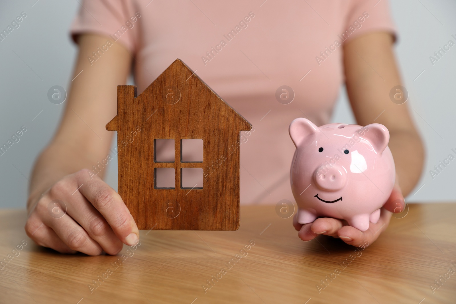 Photo of Woman holding piggy bank and house model at wooden table, closeup