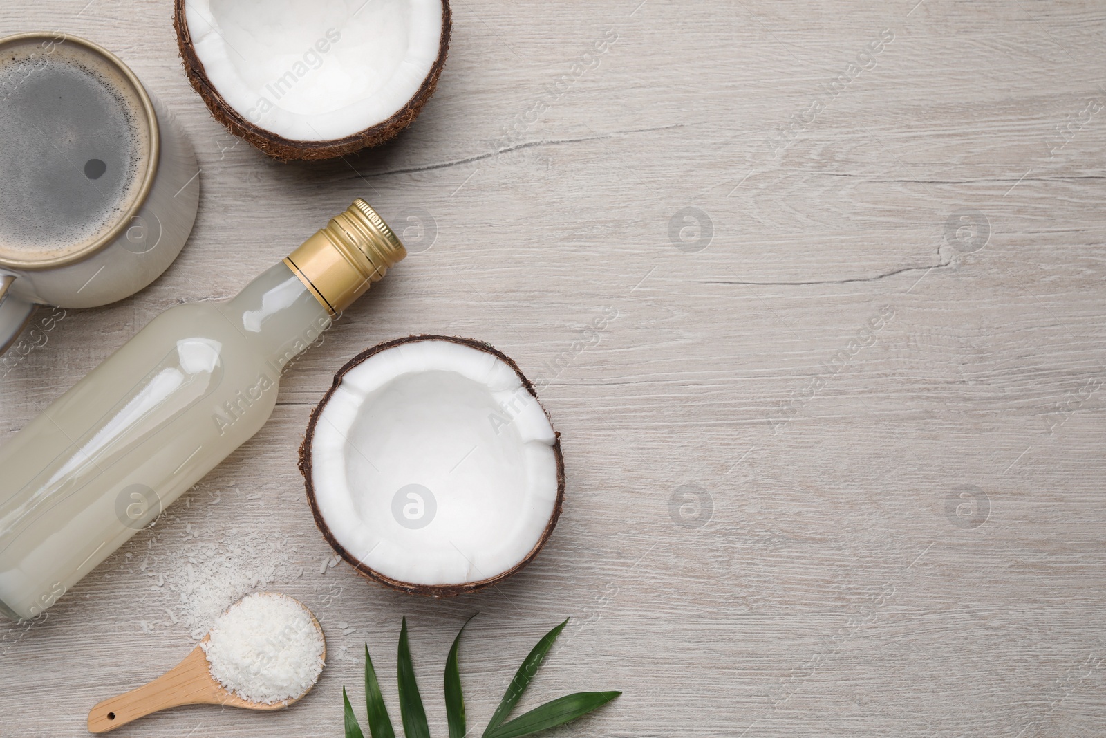 Photo of Bottle of delicious syrup, halves of coconut, flakes, cup of coffee and green leaves on white wooden table, flat lay. Space for text