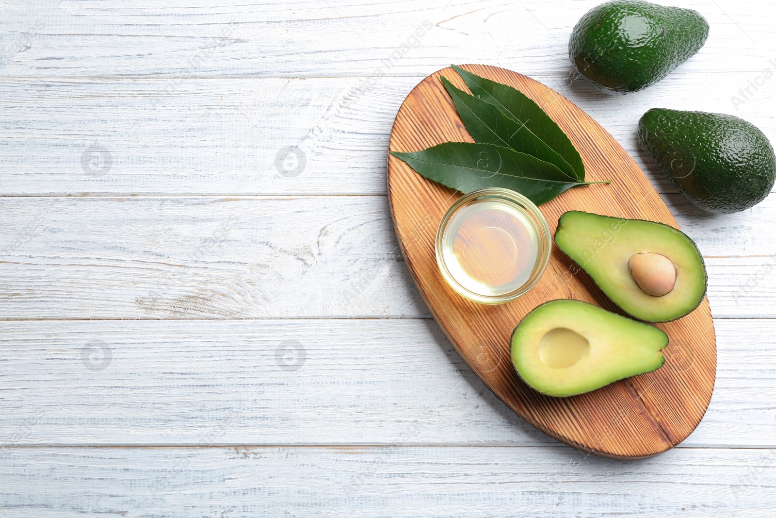 Photo of Board with bowl of natural oil and avocados on white wooden background, flat lay. Space for text