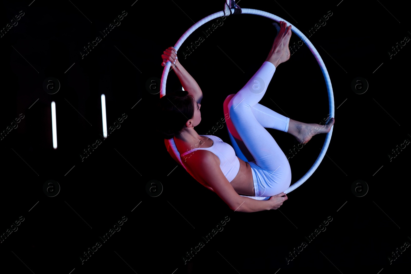 Photo of Young woman performing acrobatic element on aerial ring against dark background