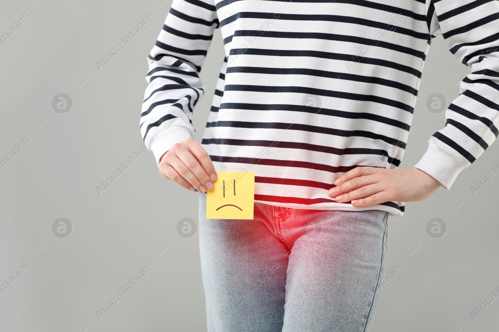 Image of Woman holding sticky note with drawn sad face and suffering from cystitis symptoms on light grey background, closeup