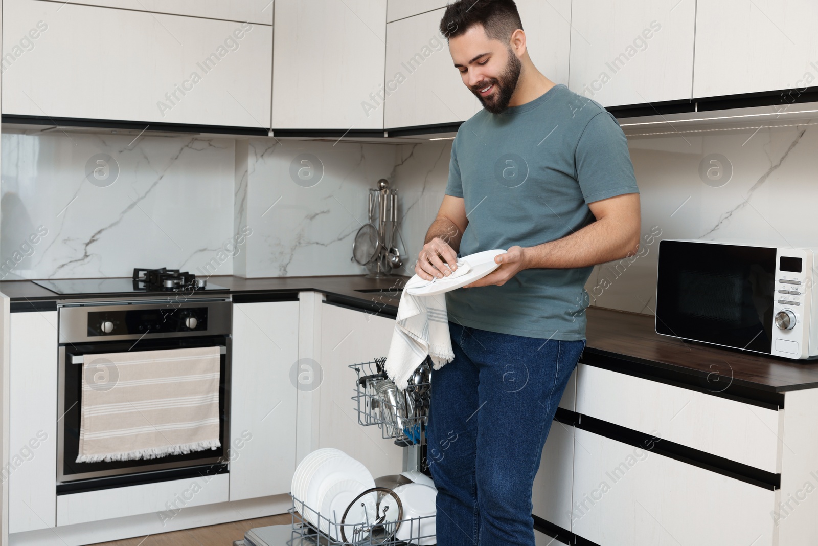 Photo of Smiling man wiping plate near open dishwasher in kitchen