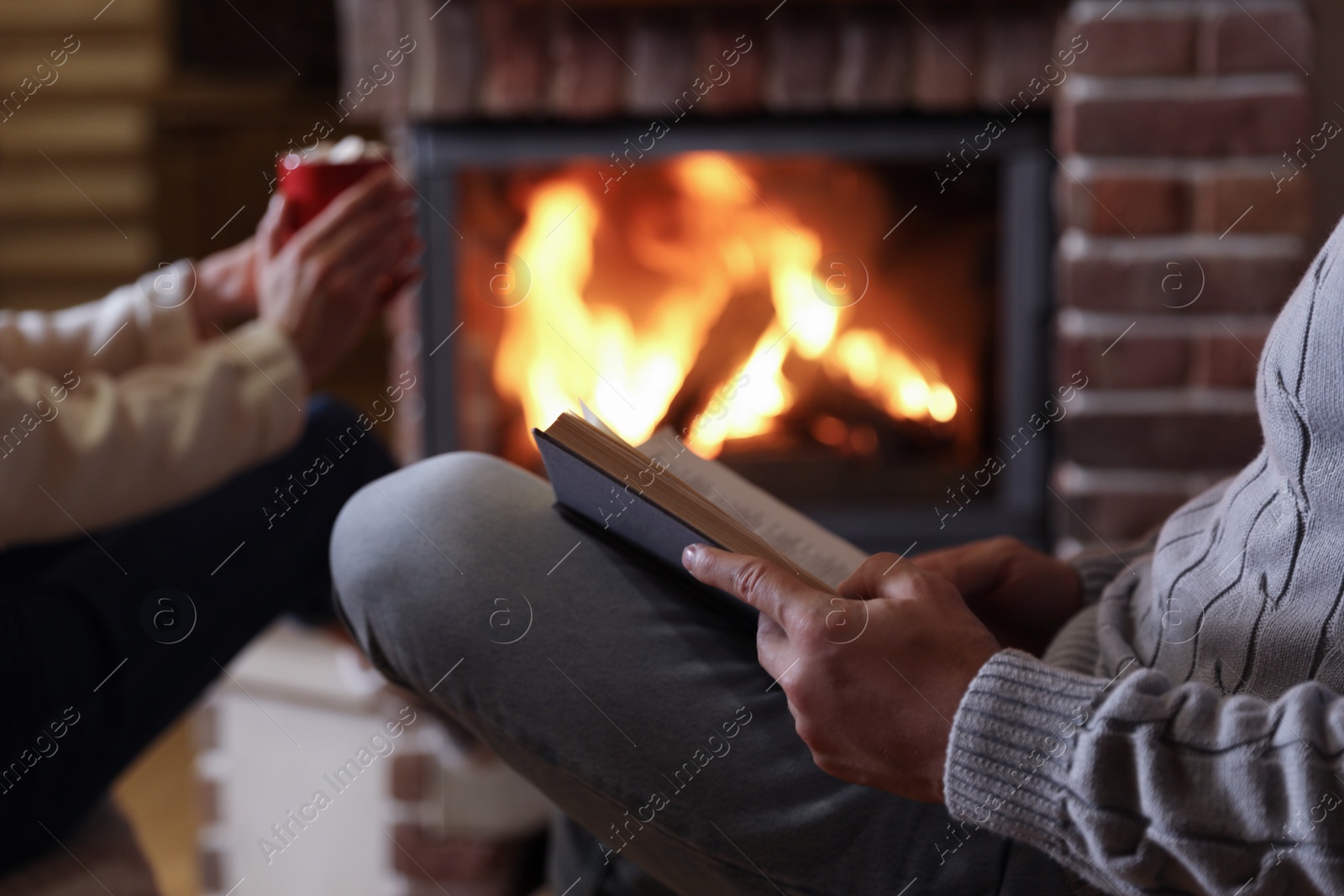 Photo of Man reading book and his girlfriend near burning fireplace at home, closeup
