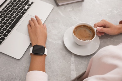 Woman with stylish smart watch and coffee working on laptop at grey table, closeup. Modern device