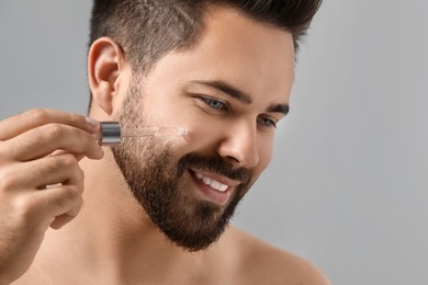 Photo of Handsome man applying cosmetic serum onto face on grey background, closeup