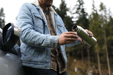 Man pouring hot drink from metallic thermos into cup lid near car, closeup