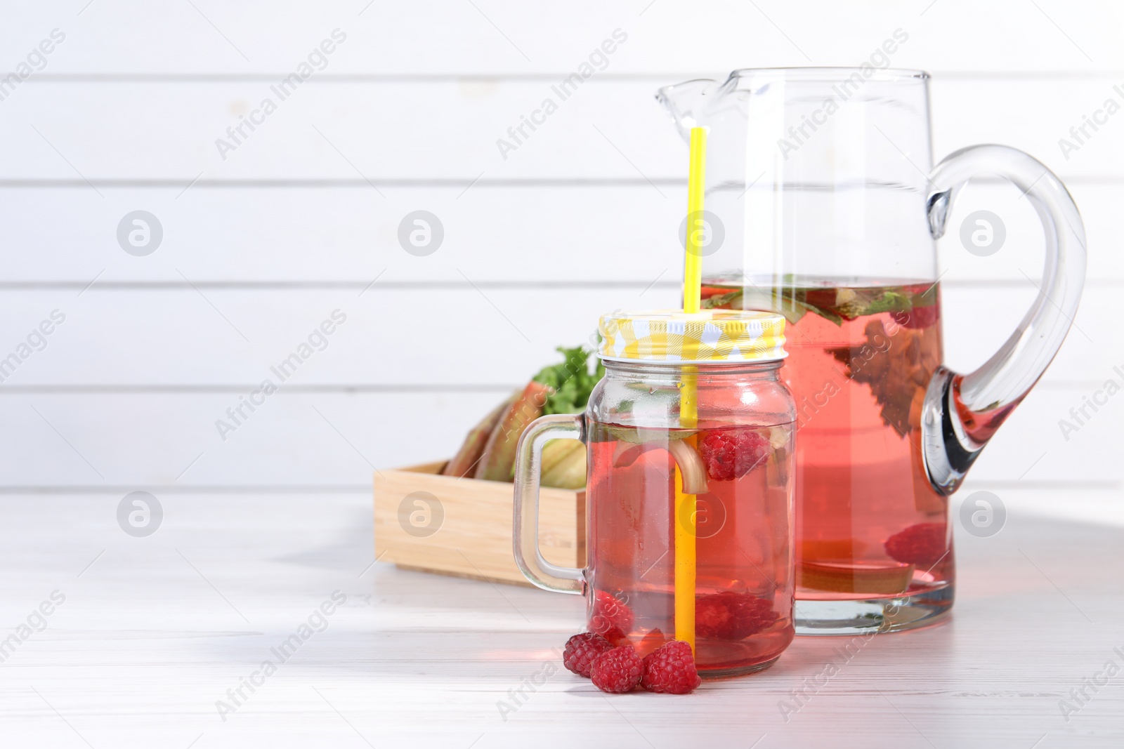 Photo of Mason jar and jug of tasty rhubarb cocktail with raspberry on white wooden table, space for text