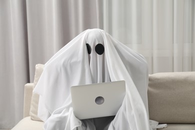 Photo of Overworked ghost. Person covered with white sheet using laptop on sofa at home