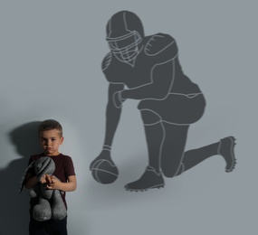Image of Little boy with soft toy dreaming to be American football player. Silhouette of man behind kid's back