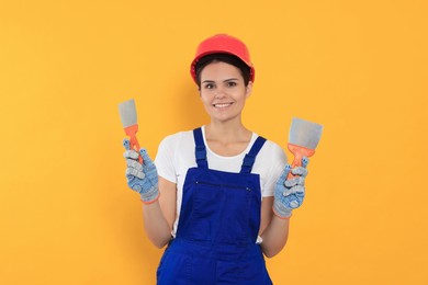 Photo of Professional worker with putty knives in hard hat on orange background