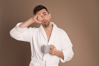 Young man in bathrobe with cup of coffee on brown background