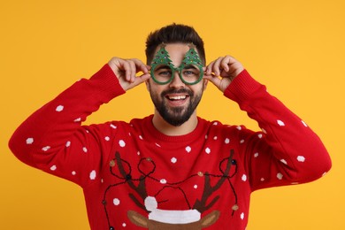 Photo of Happy young man in Christmas sweater and funny glasses on orange background