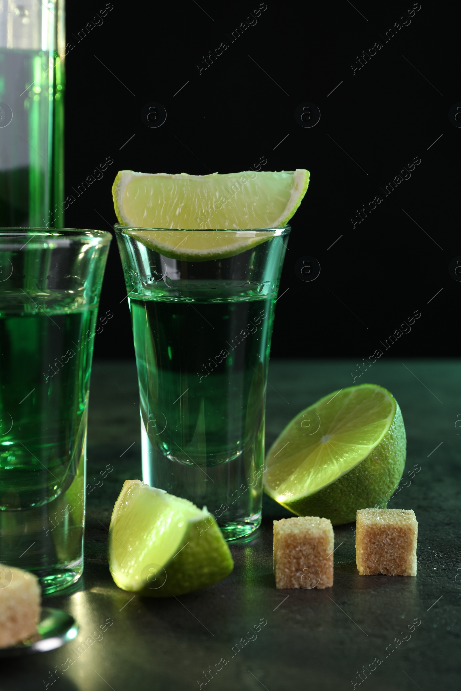 Photo of Absinthe in shot glasses, lime and brown sugar cubes on gray table against black background. Alcoholic drink