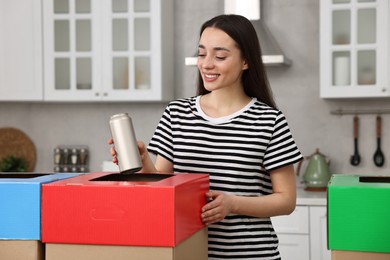 Photo of Garbage sorting. Smiling woman throwing metal can into cardboard box in kitchen