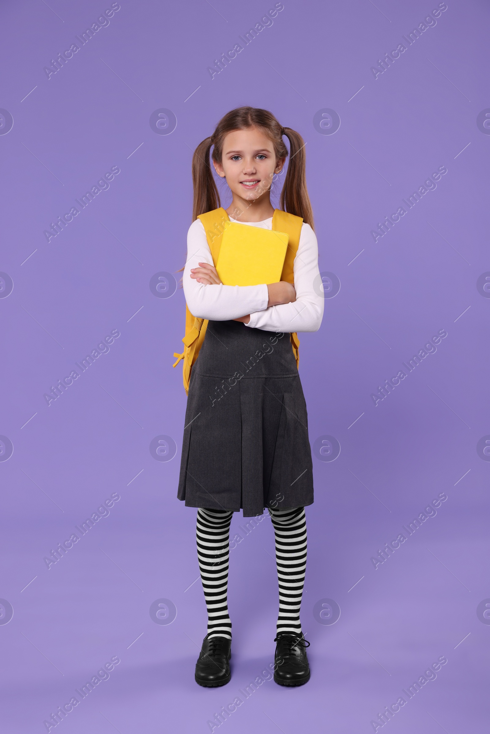 Photo of Smiling schoolgirl with backpack and book on violet background