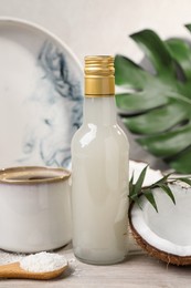 Photo of Bottle of delicious syrup, halves of coconut, flakes, cup of coffee and green leaves on white wooden table