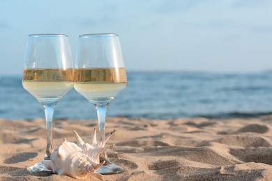 Glasses of tasty wine and seashell on sand near sea, space for text