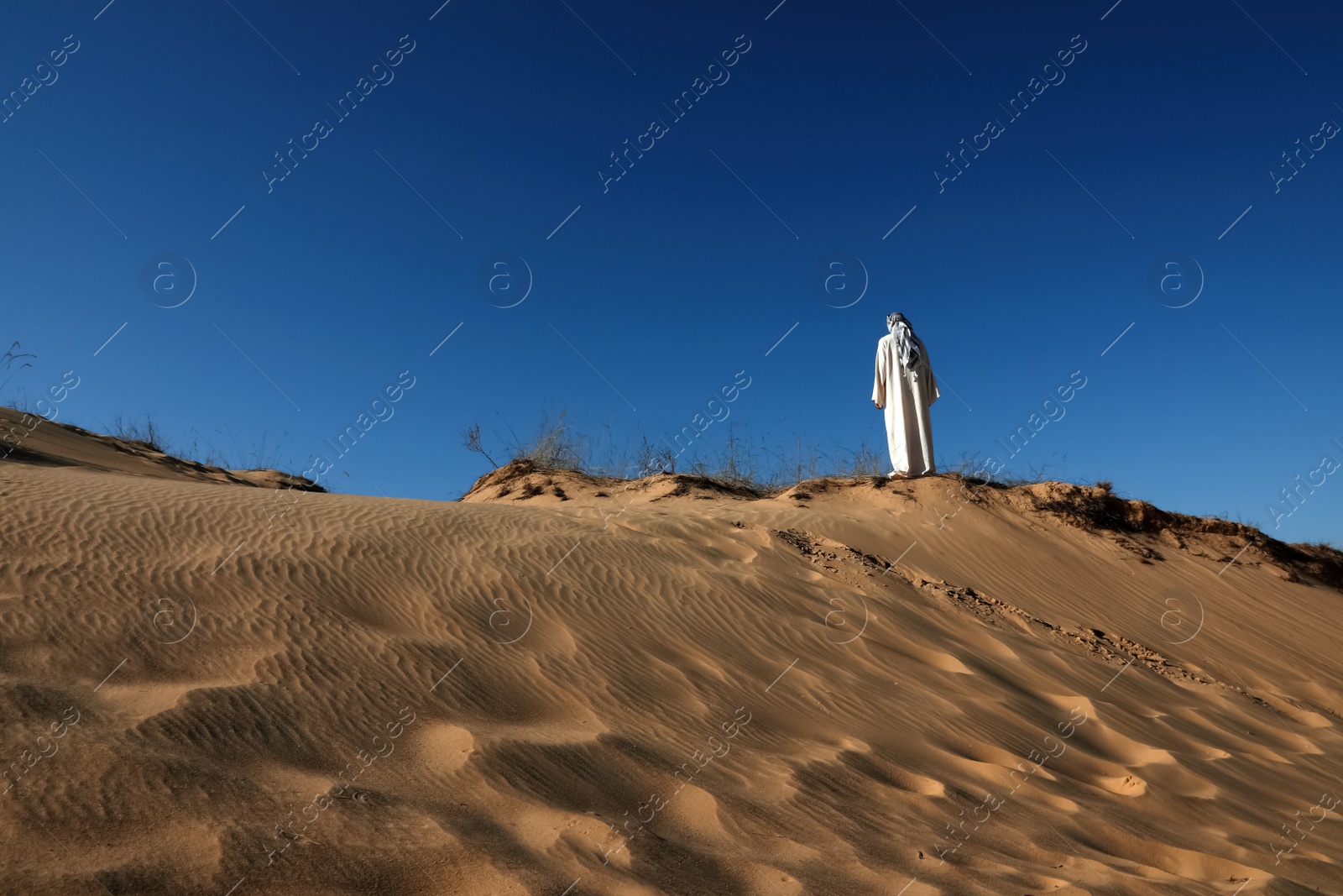 Photo of Man in arabic clothes walking through desert on sunny day, back view