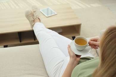 Photo of Young woman with cup of hot tea relaxing on sofa at home, closeup