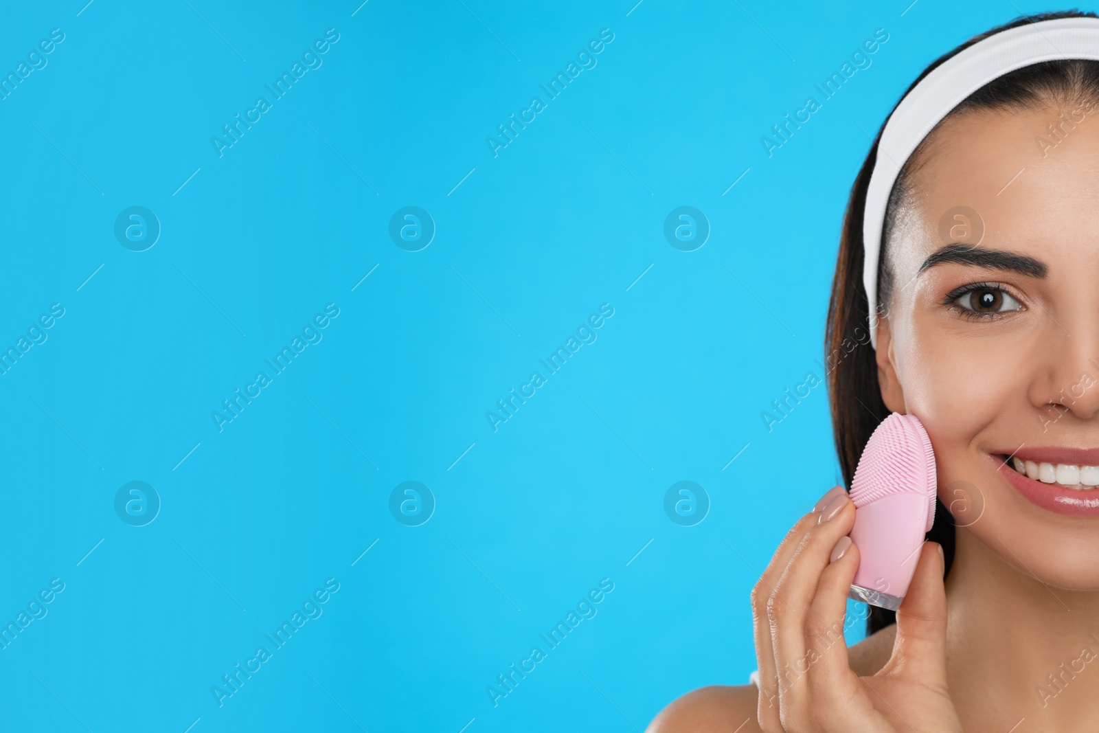 Photo of Young woman using facial cleansing brush on light blue background, space for text. Washing accessory