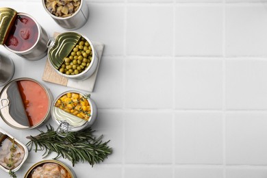 Open tin cans with different products and rosemary on white tiled table, flat lay. Space for text