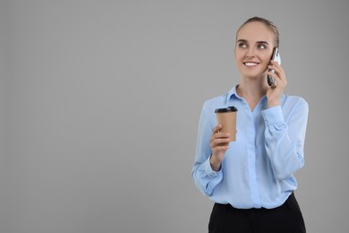 Photo of Happy young secretary with paper cup of drink talking on smartphone against grey background, space for text