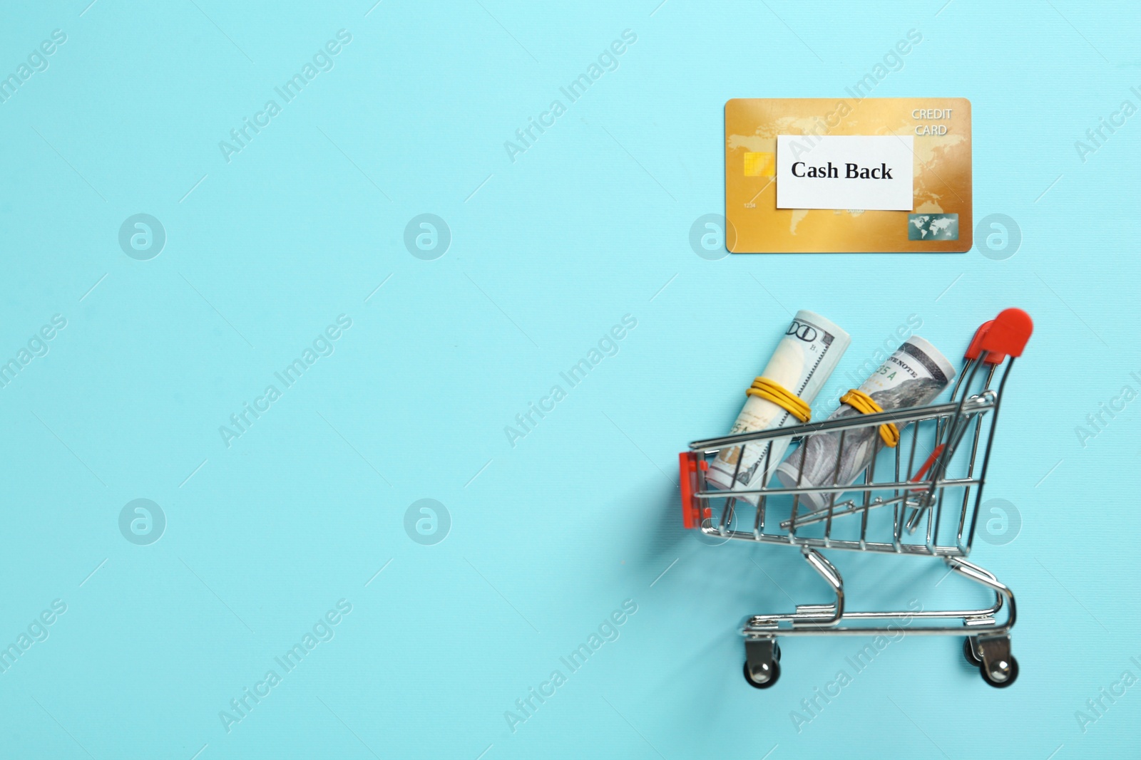 Photo of Credit card and rolled dollar banknotes in shopping cart on light blue background, flat lay with space for text. Cashback concept