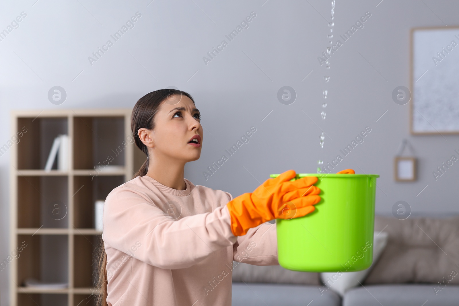 Photo of Young woman collecting leaking water from ceiling at home. Time to call roof repair service