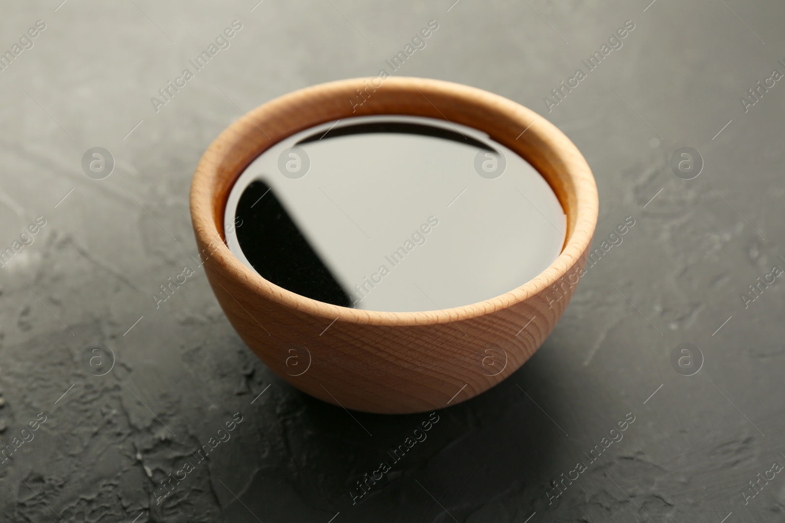 Photo of Soy sauce in wooden bowl on black textured table, closeup