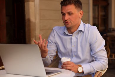 Photo of Handsome man with cup of coffee working on laptop at table in outdoor cafe